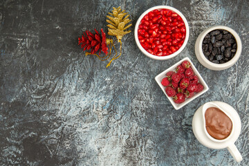 top view bowls with raspberries raisins pomegranates chocolate red and golden pinecones on grey-white background with copy space