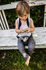 niño en camiseta de tirantes blancas sentado en un banco blanco de jardín con cámara de fotos colgada al cuello y carrete de fotos entre las manos