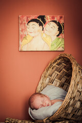 bebé recién nacido dentro de una cesta de mimbre con fondo granate y cuadro de geishas encima