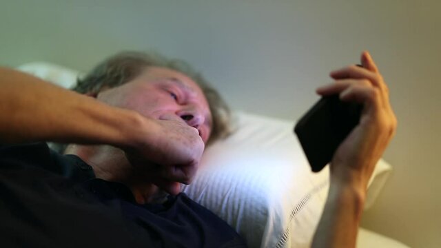 Older man lying in bed reading content online on cellphone device