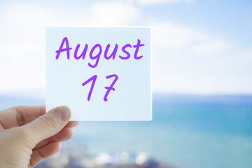 August 17th. Hand holding sticker with text August 15 on the blurred background of the sea and sky. Copy space for text. Month in calendar concept