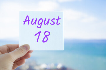 August 18th. Hand holding sticker with text August 15 on the blurred background of the sea and sky. Copy space for text. Month in calendar concept