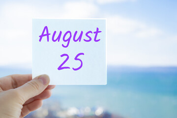 August 25th. Hand holding sticker with text August 15 on the blurred background of the sea and sky. Copy space for text. Month in calendar concept