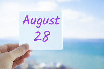 August 28th. Hand holding sticker with text August 15 on the blurred background of the sea and sky. Copy space for text. Month in calendar concept