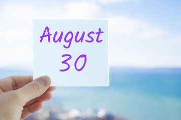 August 30th. Hand holding sticker with text August 15 on the blurred background of the sea and sky. Copy space for text. Month in calendar concept