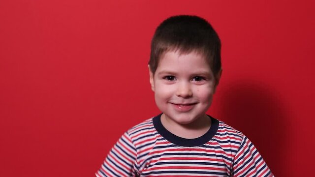 A small baby boy 3-4 years old laughs cheerfully on a red background in a striped bright T-shirt. Happy childhood and fun. Children's laughter