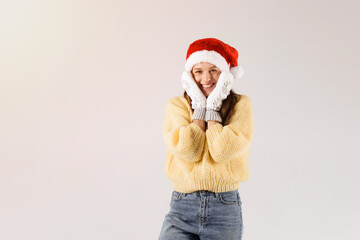 Nice happy girl in Santa Claus hat and white knitted mittens posing isolated over white background, holding hands near cheeks