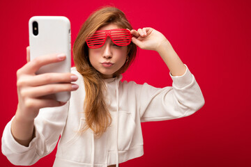 Naklejka premium Closeup portrait of attractive positive smiling young blonde woman wearing stylish white hoodie and funny colorful glasses standing isolated over colourful background holding and using mobile phone