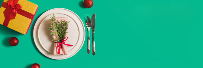 Banner with served plate and cutlery for celebration of Christmas and New Year. On plate is napkin with a Christmas tree branch, red balls. Flatlay banner on green background with balls, gift box.