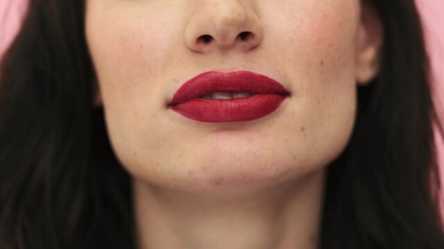 A close-up cropped view of a sexy woman with red lips is blowing bubblegum standing isolated over a pink background in the studio