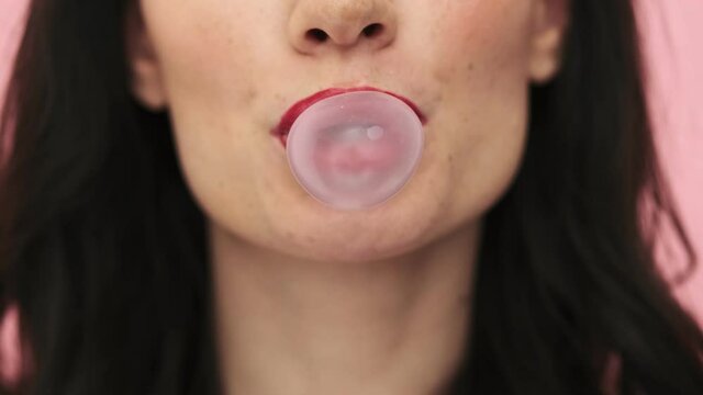 A close-up cropped view of a smiling woman with red lips is blowing bubblegum standing isolated over a pink background in the studio