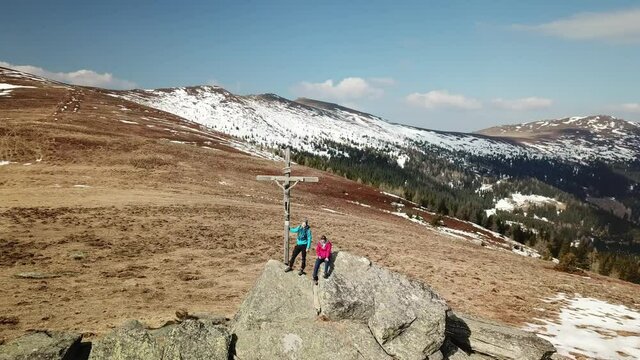 A drone capture of a couple with backpacks standing at the top of Seiner Kreuz, a peak in Austrian Alps. A wooden cross on the peak. Early spring in the mountains. Wanderlust. Achieving the goal
