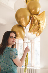 Young beautiful girl in blue dress with white polka dots celebrates her birthday and enjoys the golden balloons. Birthday alone at home during self-isolation.