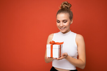 Shot of beautiful positive young blonde woman isolated over red background wall wearing white top holding gift box and looking at present