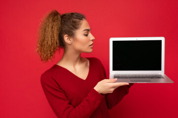 Beautiful charming fascinating young curly dark blond woman wearing red sweater standing isolated over red wall background holding computer laptop with empty copy space mock up pointing to the side