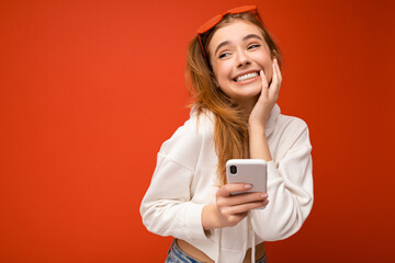 Fototapeta premium Beautiful emotional cute positive young blonde woman wearing stylish white hoodie and funny colorful glasses standing isolated over colourful background holding and using mobile phone communicating