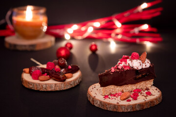 Decorated with raspberries cream, fruit and almonds, one slice of chocolate pie, wooden coaster with dates. Christmas lights, red shiny Christmas balls, black soft focus background.