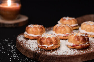Five small home baked cakes cut in halves and filled with strawberry cream, sprinkled with coconut flour on a dark wooden board. Black background, burning candle on wooden coaster
