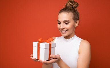 Photo shot of beautiful positive smiling young blonde woman isolated over red background wall wearing white top holding gift box and looking at present