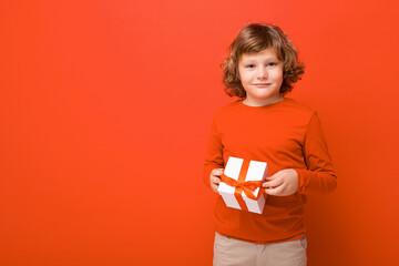 Shot of happy positive blond curly boy standing isolated over red background wall wearing red sweater holding gift box and looking at camera