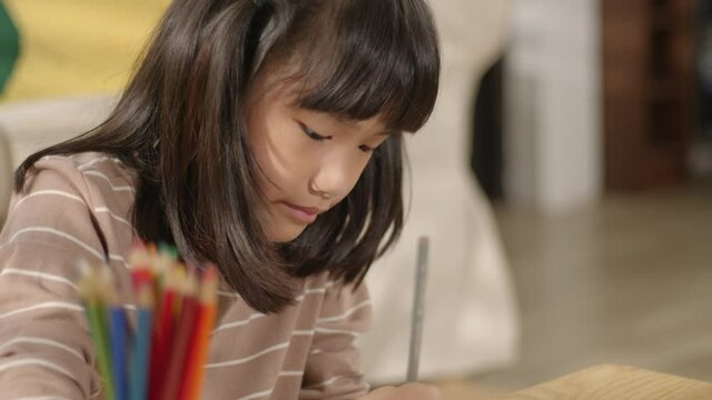 Focused Asian small kid girl drawing and painting a picture with color pencils on paper at home. Cute little daughter having fun, enjoying and learning creative art playing alone in a living room.