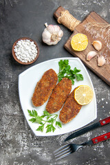 top view cutlets lemon slices parsley on white plate fork and knife garlic bowl of sea salt cutting board on grey background