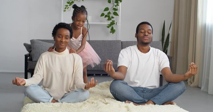 Mindful father dad and mother mom meditating sitting on floor while active energetic child daughter jumping playing, calm parents doing yoga exercise at home for stress relief relaxing with little kid