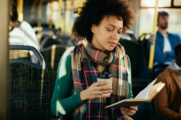Black woman reading a novel and drinking takeaway coffee while traveling by bus.