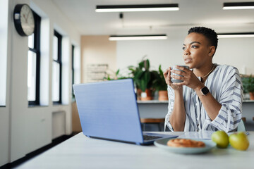 Thoughtful young businesswoman drinking coffee at laptop in office