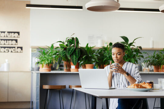 Businesswoman with coffee working at laptop in morning office lounge