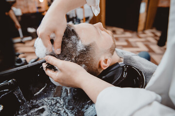 Barber massages head of client man, washes his hair with shampoo, black sink, vintage color