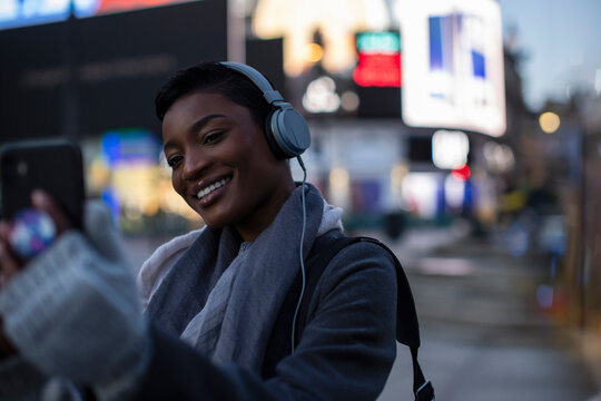 Happy young woman with headphones taking selfie in city at night