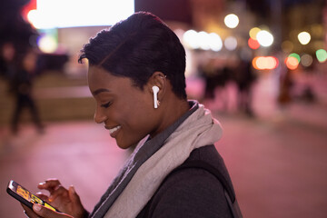 Young woman with earbud headphones using smart phone in city at night