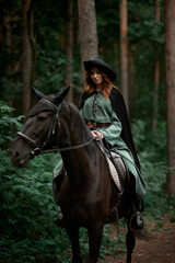 Fototapeta premium Young beautiful woman in a suit, green dress, corset, black topm hat on a black horse on nature in the forest. fairy tale, creative photo session of a girl with a horse