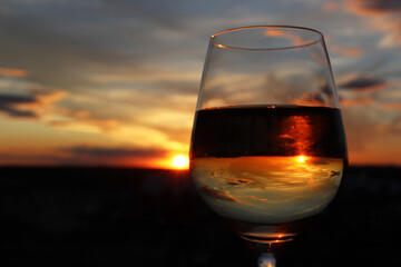 Glass with alcohol drink on beautiful sunset background, sun and sky are reflected in liquid. Concept of celebration, evening party at resort, romantic dinner outdoors