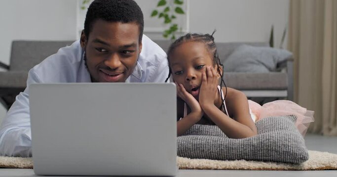 Young black man and little girl afro american father with curious emotional daughter looking at laptop lying on floor at home enjoy watching movie video online learning to work on laptop making order