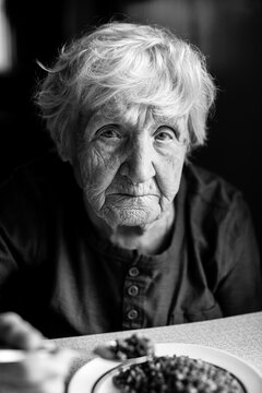 Portrait of old woman eats buckwheat porridge sitting at the table in his home. Black and white photo.