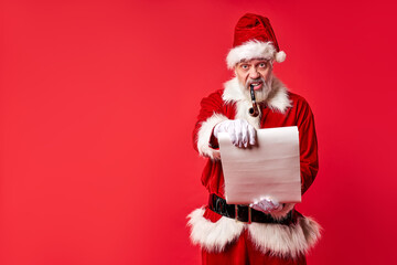 santa claus with smoking pipe in mouth, holding paper in hands isolated over red background