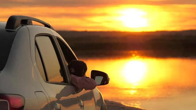A free woman traveler admires the scenery from the car window, enjoying beautiful sunrise over the lake. The girl driver stopped at the campsite in her car, looking at the sunset.