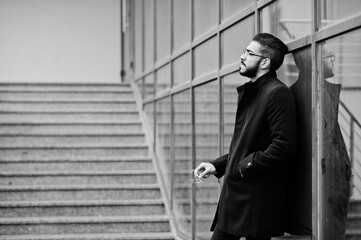 Fototapeta na wymiar Middle eastern entrepreneur wear black coat and blue shirt, eyeglasses against office building smoking cigarette.
