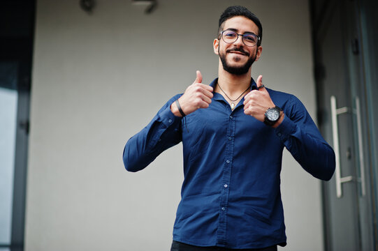 Middle eastern entrepreneur wear blue shirt, eyeglasses against office building and show thumbs up.