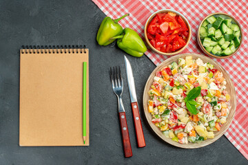 top view of veggie salad with red napkin under it and vegetables cutleries and notepad on side on dark grey background