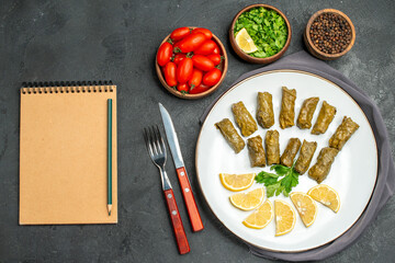 top view stuffed grape leaves on white oval plate bowls with cherry tomatoes parsley black pepper knife and fork purple kitchen towel and a notebook on dark background
