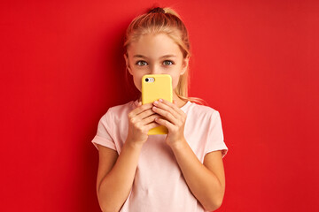 blond child girl use smartphone, isolated on red background in studio. checking news, surf the net, look at camera, closing face with yellow mobile phone