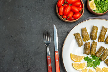 top view stuffed grape leaves lemon half slices on oval plate bowls with cherry tomatoes parsley black pepper knife and fork and purple kitchen towel on dark background with copy space