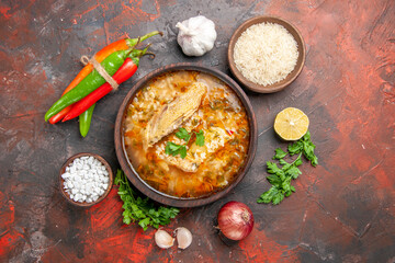 top view homemade chicken and rice soup in a bowl hot peppers bowls with sea salt and rice garlic parsley leaves red onion on dark red background with copy space