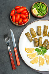 top view stuffed grape leaves and lemon half slices on white oval plate bowls with cherry tomatoes parsley knife and fork and purple kitchen towel on dark background