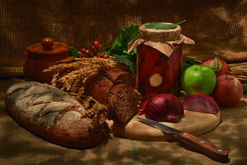 green and red apples, black bread and ears of wheat on sacking, canned red peppers in a glass jar, red onions cut with a knife with a wooden handle lies on a wooden board