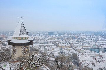 The famous clock tower on Schlossberg hill and historic buildings rooftops with snow, in Graz, Styria region, Austria, in winter day