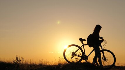 Fototapeta na wymiar A free girl travels on bike, rests, looks at the sunset and enjoys the sun. Adventure and travel concept. A healthy young tourist walks with bicycles across the field, enjoying the nature, fresh air.